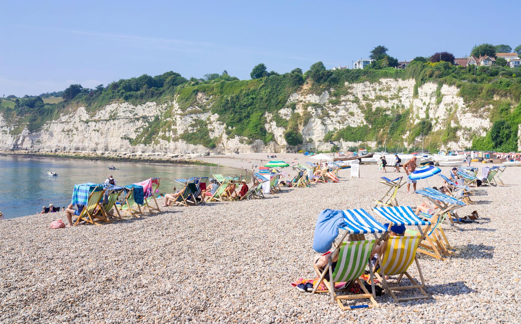 Beachgoers relax in deck chairs on a pebble beach below cliffs.
