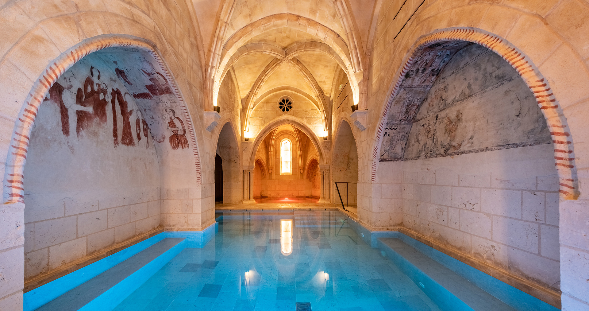 Indoor pool in a stone building with arched alcoves containing faded murals.