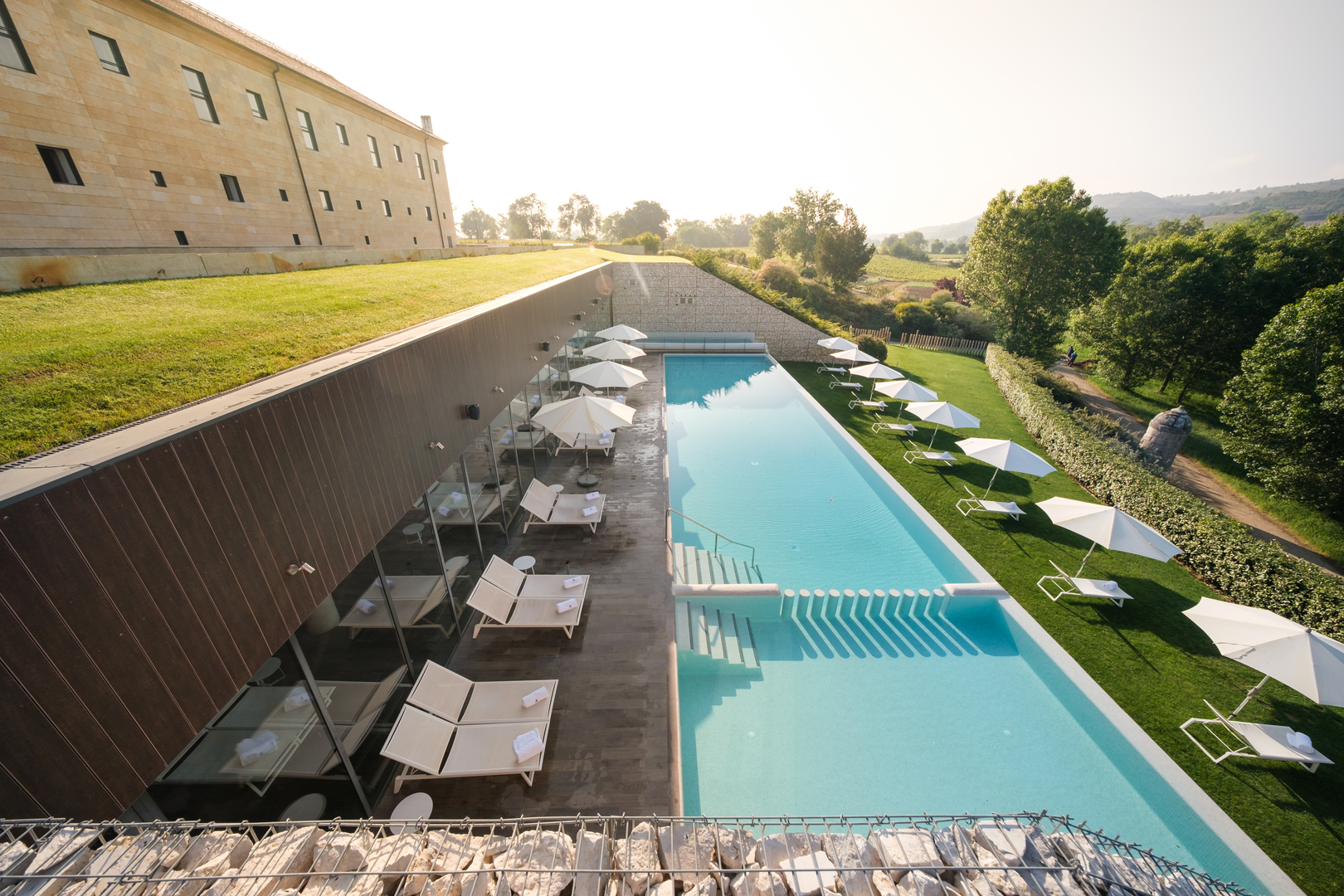 Aerial view of a long, light blue pool at Castilla Termal Monasterio de Valbuena. Lounge chairs and umbrellas are positioned around the pool.