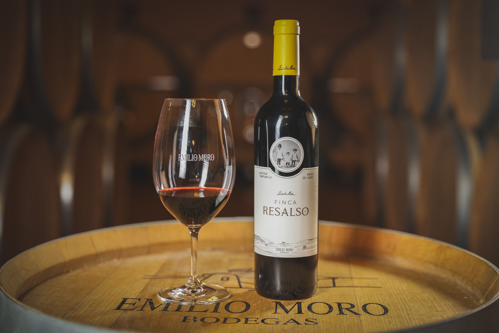 Bottle of Emilio Moro Finca Resalso wine and a glass of red wine on a wine barrel.