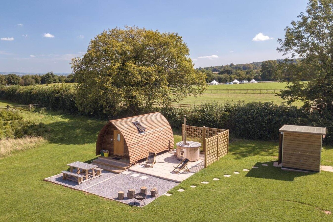 Aerial view of a wooden cabin with a hot tub and picnic table, situated in a grassy field.